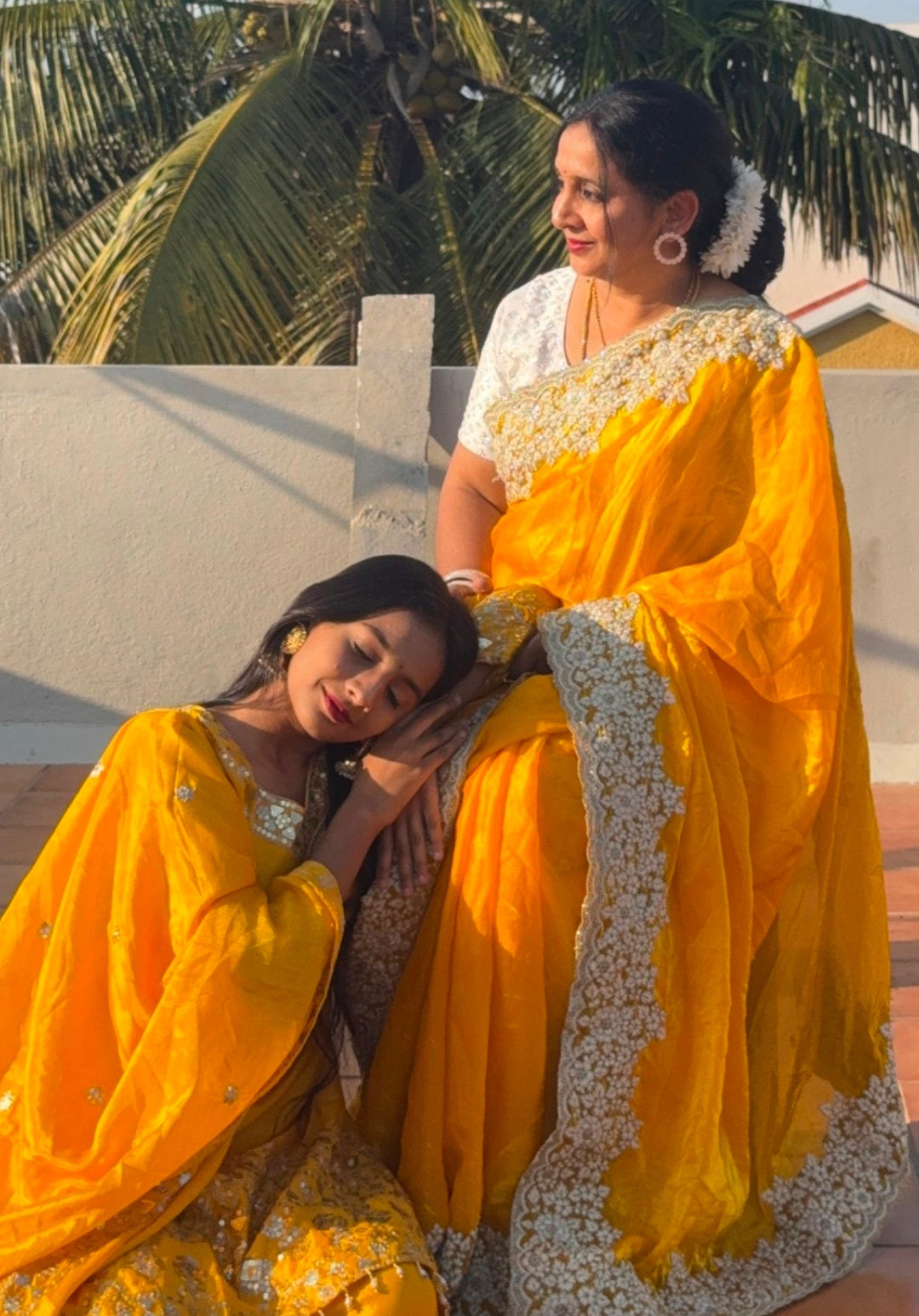 Two women in traditional yellow and white sarees sitting on a rooftop with palm trees in the background.